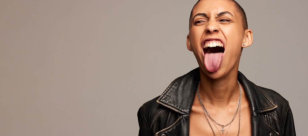 Excited androgynous woman sticking out her tongue on gray background. Expressive woman with shaved head looking away at copy space.