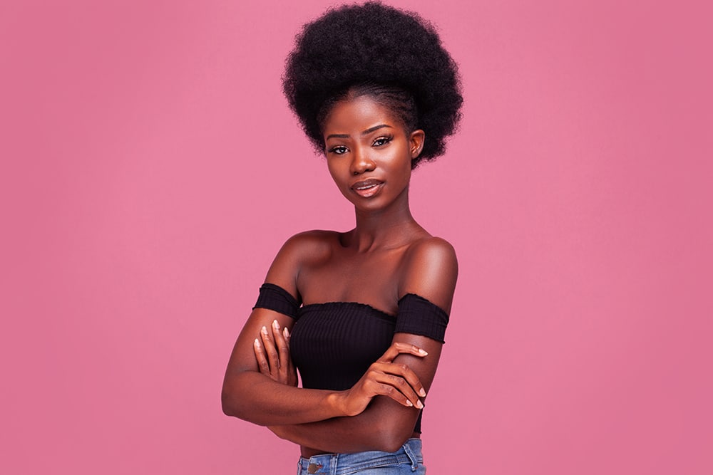 Confident African American girl with afro hair standing light smiling with arms folded in black bare shoulder top and blue denim jeans isolated on pink background.
