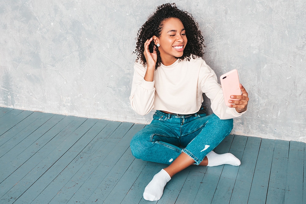 Beautiful black woman with afro curls hairstyle.Smiling model in white trendy sweater. Sexy carefree female sitting near gray wall in studio interior.She taking selfie photos for instagram