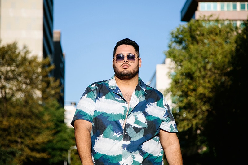 Fat latin man with sunglasses, walking, with a serious expression. With a street in the background. Latins, plus side and curvy models concept. Copyspace