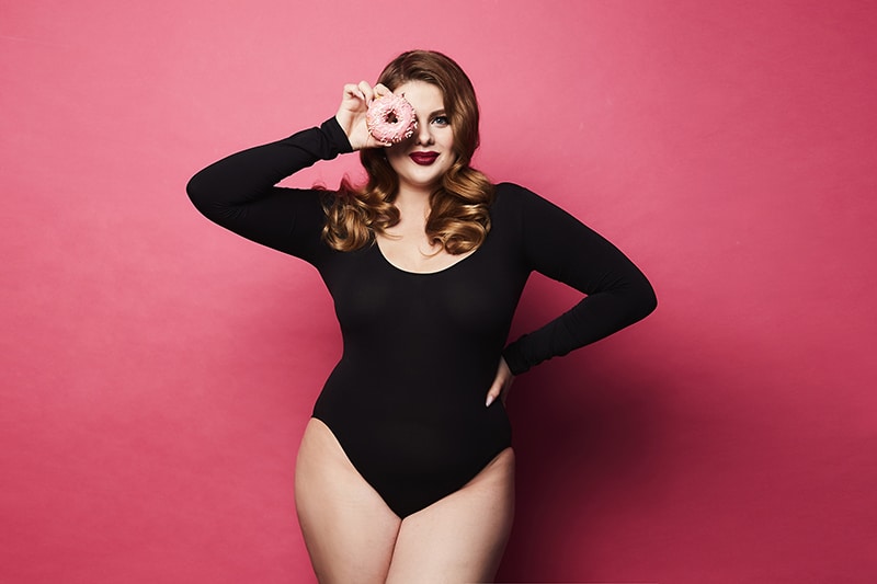 Plus size sexy model girl, fashionable blonde in black bodysuit, with bright makeup and with donut in her hand smiling and posing at pink background in studio