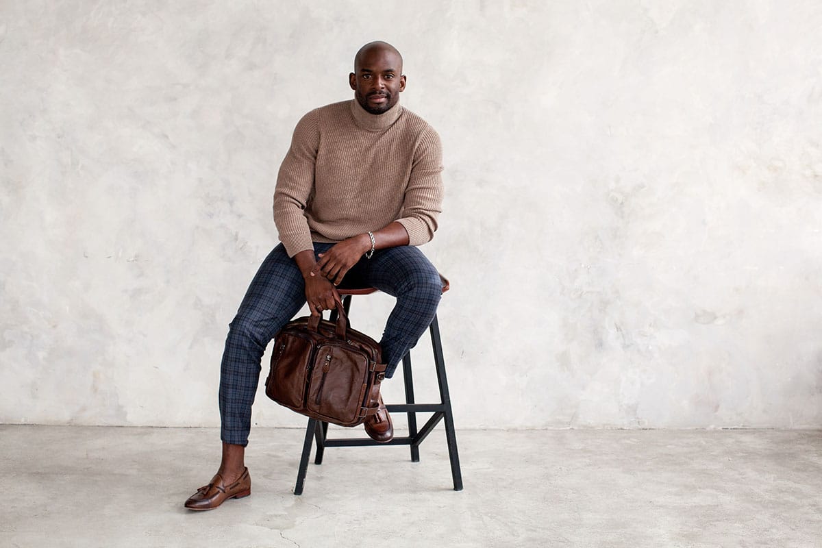 Stylish man sitting on chair and looking camera. Confident fashion model in fashionable outfit turtleneck knitted sweater, plaid trousers, leather shoes and bag. Office clothes style for businessmen